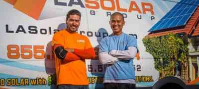 Two smiling employees in orange and blue shirts stand confidently in front of an LA Solar Group branded van, highlighting their commercial solar services.